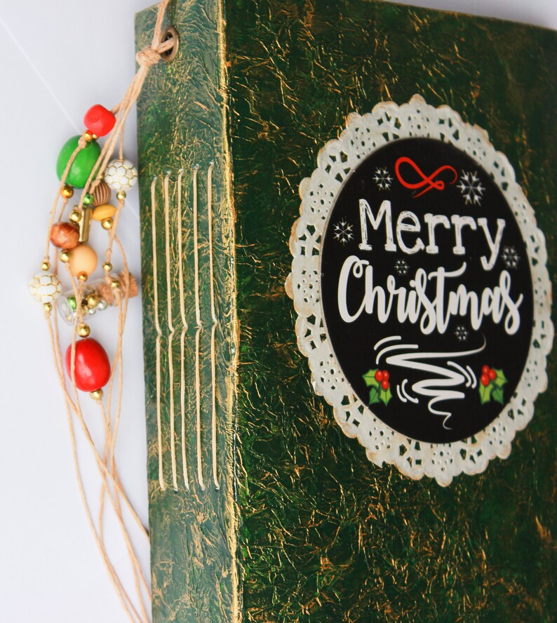 May include: A green journal with gold accents and a white lace doily with the text "Merry Christmas" in black lettering.