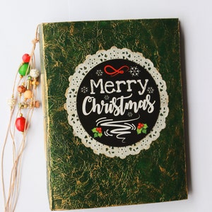 May include: A green, textured box with a white doily and a black circle that says "Merry Christmas" in white lettering. The box has a brown string with colorful beads attached.