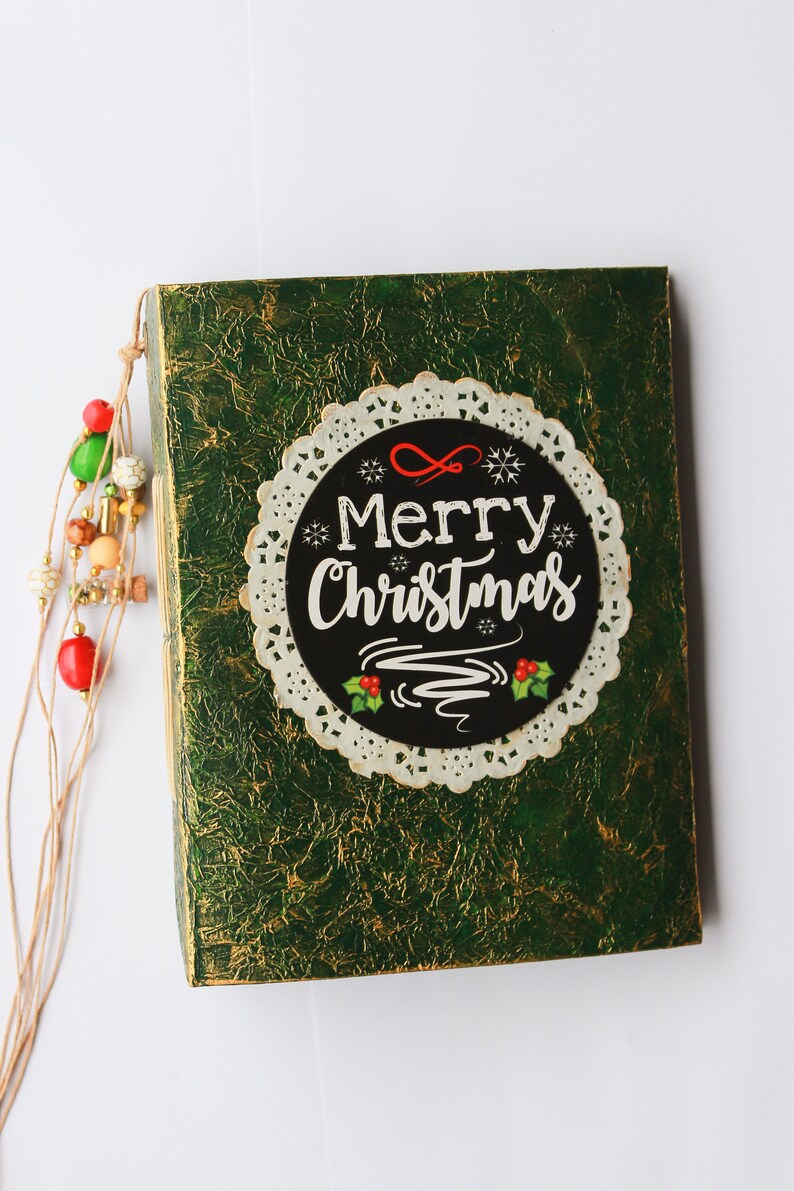 May include: A green, textured box with a white doily and a black circle that says "Merry Christmas" in white lettering. The box has a brown string with colorful beads attached.