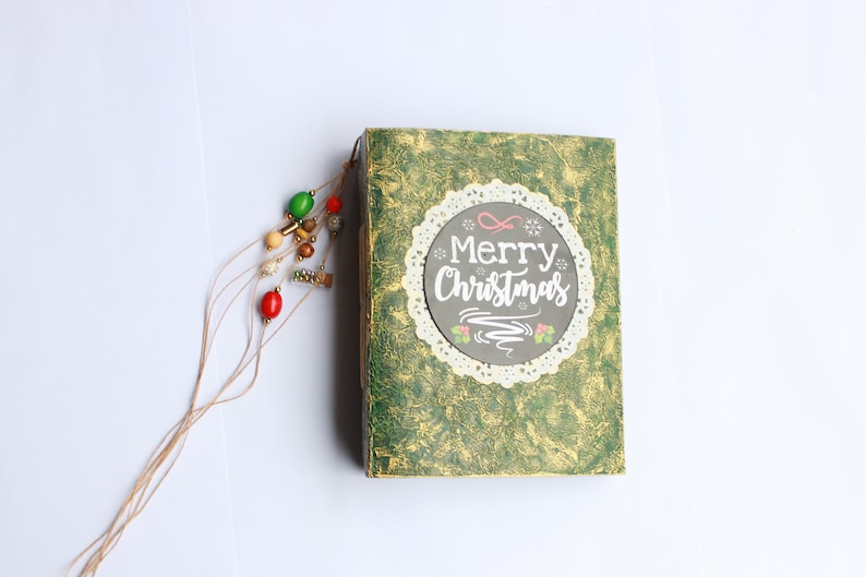 May include: A green journal with a gold and white design. The cover has a white lace circle with the text "Merry Christmas" in white lettering. A brown string with colorful beads is attached to the journal.