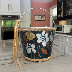 May include: A decorative bucket featuring a dark teal background with a floral pattern of white and gold flowers. The rim and base are trimmed with gold braided cord. A silver metal handle is attached, adorned with a long, tan velvet ribbon.