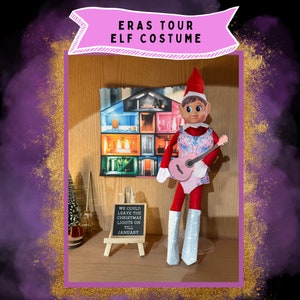 Peut inclure: Une poupée d'elfe portant un costume pailleté rose et bleu, tenant une guitare rose. L'elfe est debout sur une étagère avec une pancarte qui dit "We could leave the Christmas lights on till January".