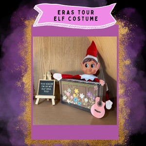 May include: An elf doll dressed in a red and white costume is playing a pink toy guitar. The elf is sitting on a box with a floral design. A small chalkboard sign reads "You know I'm in my elf era, too".