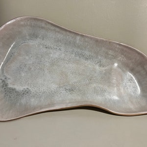 May include: A large, white ceramic serving dish with a slightly irregular, organic shape. The dish has a smooth, glossy finish and a subtle, speckled texture.