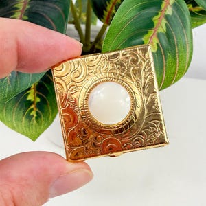 Puede incluir: Un estuche cuadrado dorado con un diseño ornamental y arremolinado. El centro presenta una piedra redonda blanca, similar a una perla. El estuche se sostiene en una mano, con follaje verde en el fondo.