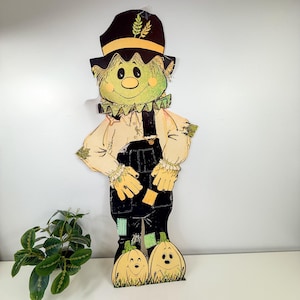 May include: A flat, cutout-style scarecrow decoration featuring a green face, a black hat with yellow wheat detail, a cream-colored shirt, and black overalls. The scarecrow stands atop two smiling, light-colored pumpkins with green stems.