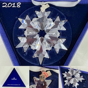 May include: A clear, faceted crystal snowflake ornament with a silver-toned metal tag engraved with the year 2018. The ornament is suspended by a white ribbon and presented in a dark blue triangular box labeled Annual Edition Ornament 2018.
