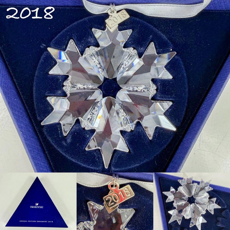 May include: A clear, faceted crystal snowflake ornament with a silver-toned metal tag engraved with the year 2018. The ornament is suspended by a white ribbon and presented in a dark blue triangular box labeled Annual Edition Ornament 2018.