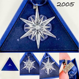 May include: A clear, multi-pointed crystal star ornament featuring a faceted design. It includes a blue ribbon for hanging and a small silver-toned metal tag engraved with the year 2005. The ornament is presented in a triangular blue box.
