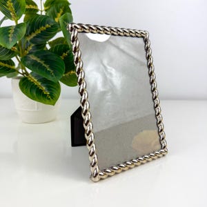 May include: A rectangular picture frame with a metallic silver finish and a twisted rope-style border. The frame features a clear protective front panel and a black easel-style stand attached to the back for upright display on a flat surface.