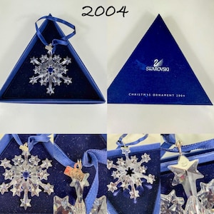 May include: A clear crystal snowflake ornament with a blue ribbon, featuring a metal tag engraved with 2004. It is presented in a triangular blue box with the Swarovski logo and the text Christmas Ornament 2004 printed on the lid.