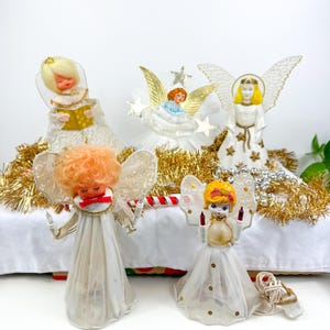 Op de afbeelding: Een verzameling vintage kerstboomtoppers in de vorm van engelen. De engelen hebben witte jurken, vleugels en verschillende haarkleuren. Sommigen houden kaarsen of boeken vast en zijn versierd met gouden en zilveren accenten. De scène staat op een wit oppervlak met gouden slingers.