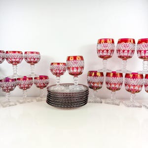 以下が含まれることがあります： ルビーレッドとゴールドリムのクリスタルガラス製品のコレクション。ワイングラスと小皿が含まれます。グラスはカットパターンデザインで、白い表面に配置されています。