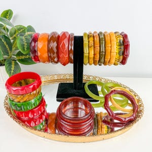 May include: A collection of resin bangle bracelets in various colours including red, orange, brown, yellow, and green. Some bracelets feature faceted geometric patterns, while others have a smooth, bamboo-like texture, displayed on a black stand.