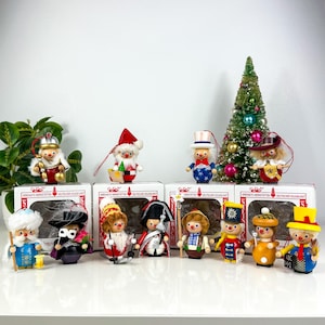 May include: A collection of vintage wooden Christmas ornaments, including Santa Claus figures, a snowman, and other characters. The ornaments are displayed on white boxes and in front of a small decorated Christmas tree. The ornaments are painted in bright colors.