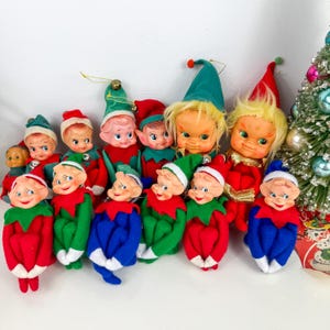 May include: A collection of twelve vintage-style elf dolls with painted faces, pointed ears, and felt clothing in red, green, and blue. Each doll wears a matching pointed hat, some with small bells attached, and features soft hair and posable limbs.