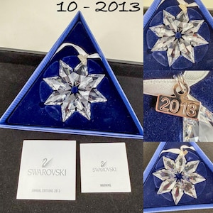 May include: A clear faceted crystal star ornament with a white ribbon and a silver-toned metal tag engraved with the year 2013. The ornament is presented in a triangular blue box with two white informational cards labeled Swarovski Annual Editions 2013.