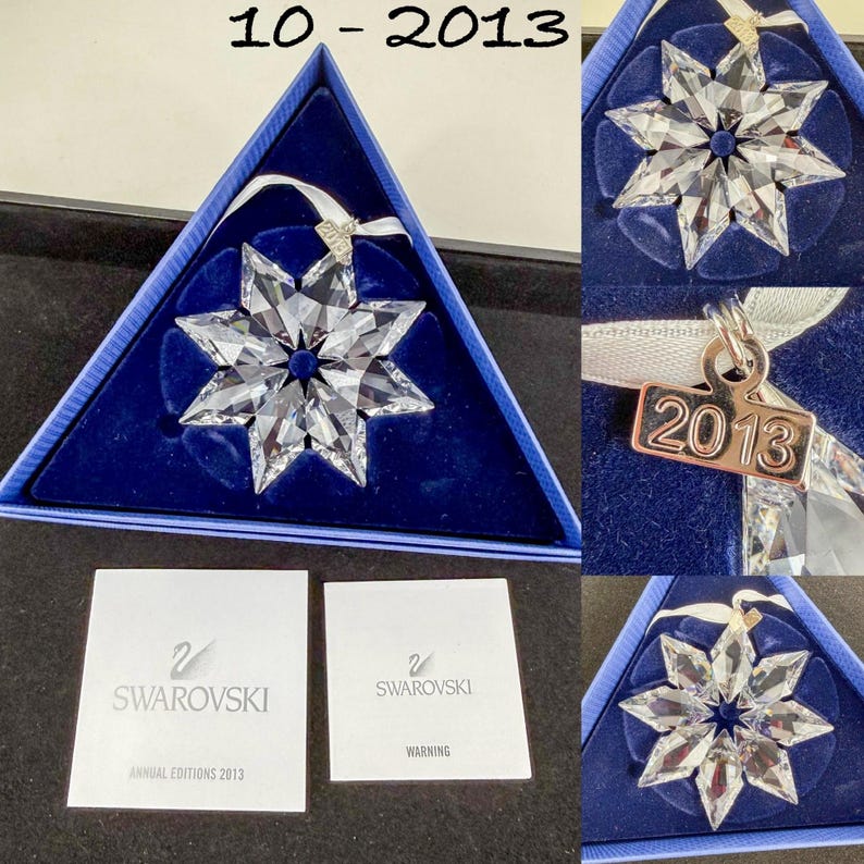 May include: A clear faceted crystal star ornament with a white ribbon and a silver-toned metal tag engraved with the year 2013. The ornament is presented in a triangular blue box with two white informational cards labeled Swarovski Annual Editions 2013.