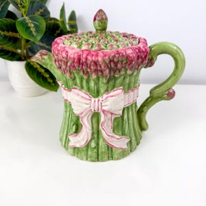 May include: A ceramic teapot shaped like a bundle of asparagus with green stalks, a pink-tipped lid, and a decorative white ribbon with pink trim tied around the center. The handle is curved and green, featuring a small bud at the base.