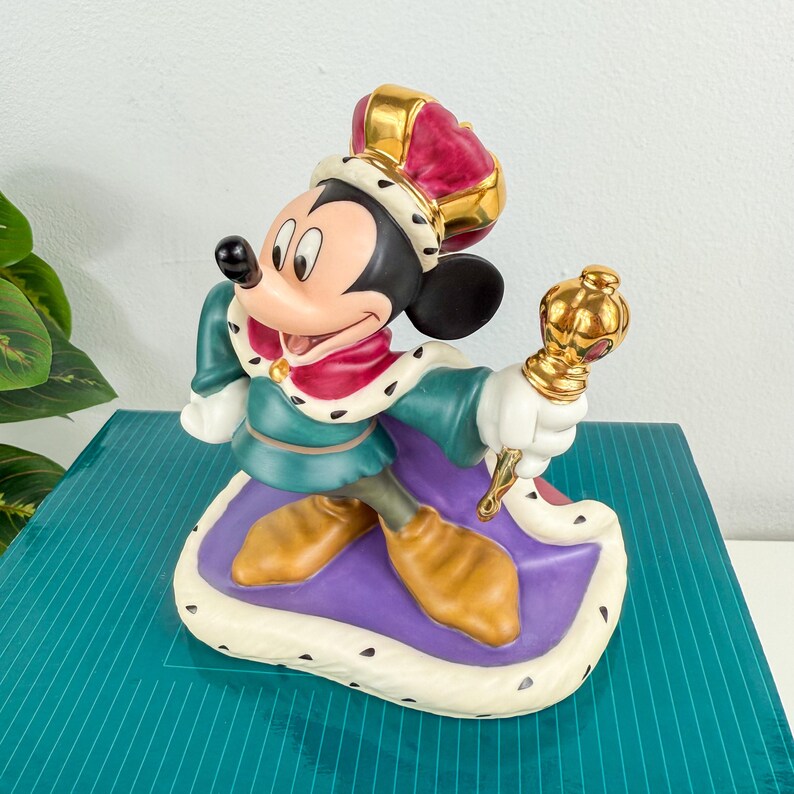 Op de afbeelding: Een keramieken beeldje van Mickey Mouse gekleed als koning, met een gouden kroon met een bordeauxrode top, een groene tuniek en een paarse cape. Hij houdt een gouden scepter vast en staat op een paarse en witte basis.