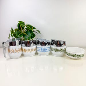 Könnte beinhalten: Set aus vier Vintage-Glas-Zucker- und Sahne-Spendern mit verchromten Deckeln. Die Spender haben Blumenmuster und die Wörter "Cream", "Sugar" und "Sugar" sind darauf gedruckt. Eine passende grüne und weiße Schüssel ist ebenfalls enthalten.
