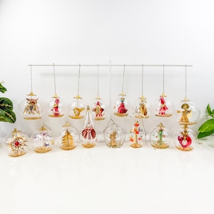 May include: A collection of vintage Christmas ornaments. Clear glass globes feature various designs, including angels, trees, and stars, with gold and red accents. Each ornament hangs from a thin wire, creating a festive display.