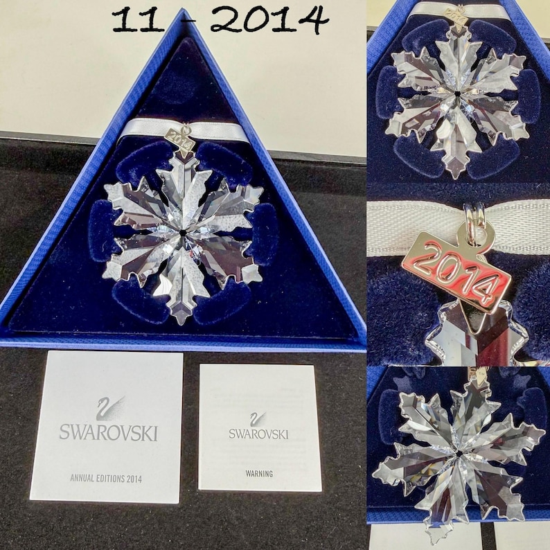 May include: A clear crystal snowflake ornament with a white ribbon and a silver-toned metal tag engraved with 2014. The ornament is housed in a triangular blue velvet-lined box and includes two white informational booklets with the Swarovski logo.