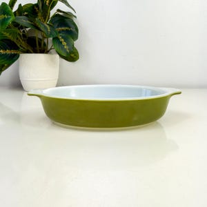 May include: An oval-shaped baking dish with a smooth, olive green exterior and a glossy white interior. The dish features two small, integrated handles on opposite sides, providing a simple, functional design for oven-to-table use.
