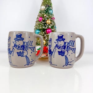 May include: Two grey ceramic mugs with blue snowman designs. Each mug features a snowman wearing a top hat and scarf, holding a broom and a book. A small Christmas tree with colourful baubles is in the background.
