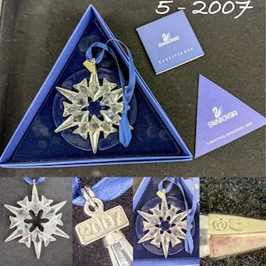 May include: A clear faceted crystal snowflake ornament with a blue ribbon and a metal tag engraved with 2007. The set includes a triangular blue presentation box, a certificate of authenticity, and a triangular card labeled Swarovski Christmas Ornament 2007.