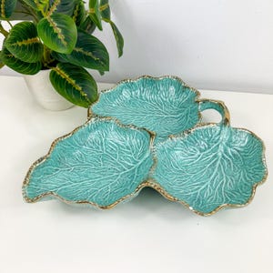 May include: A turquoise ceramic serving dish in the shape of three overlapping leaves. The dish has a gold trim and detailed vein patterns. A handle is on the top leaf. The dish is on a white surface with a potted plant in the background.