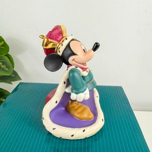 Op de afbeelding: Een keramieken beeldje van Mickey Mouse als koning. De figuur draagt een rode en gouden kroon, een groene en gouden tuniek en een paarse en witte mantel. Mickey heeft een zwarte neus en oren en een glimlachende uitdrukking.
