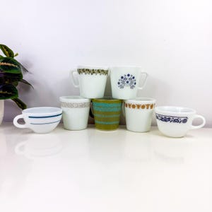 Puede incluir: Seis tazas de cerámica blanca con diferentes diseños de colores y patrones alrededor del borde. Las tazas están dispuestas en semicírculo sobre una superficie blanca.