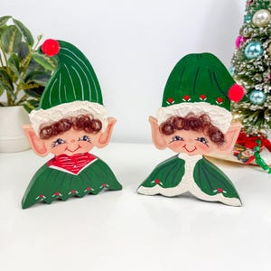 Op de afbeelding: Twee houten kerstelfjes. Elke elf heeft een groene hoed met een rode pompon, een wit omrand gezicht en een groene outfit met een rode kraag. De elfen hebben geschilderde gezichten met roze wangen en lachende uitdrukkingen. Op de achtergrond staat een kleine kerstboom.