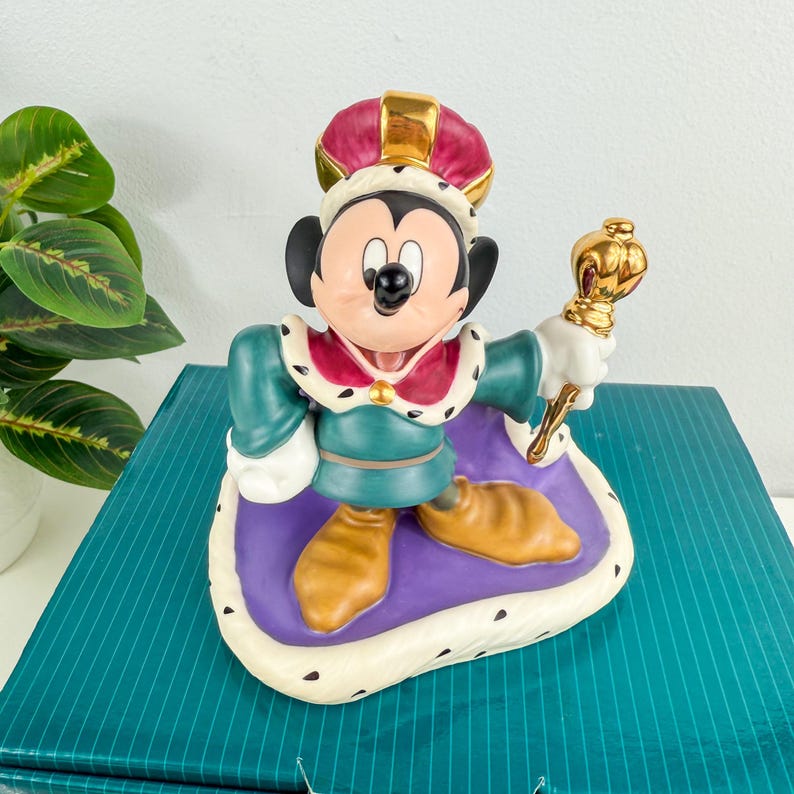Op de afbeelding: Een keramieken beeldje van Mickey Mouse als koning, met een rode en gouden kroon en een gouden scepter. Hij draagt een groene tuniek, een paarse cape en bruine schoenen. Het beeldje staat op een witte en paarse basis.