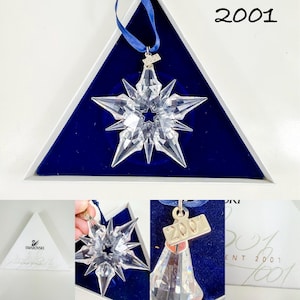 May include: A clear, multi-pointed crystal star ornament featuring a central star cutout. It includes a blue ribbon for hanging and a small metal tag engraved with the year 2001. The ornament is presented in a triangular white box with blue velvet.