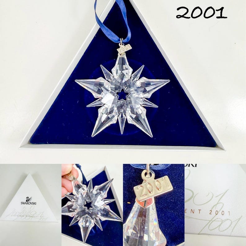 May include: A clear, multi-pointed crystal star ornament featuring a central star cutout. It includes a blue ribbon for hanging and a small metal tag engraved with the year 2001. The ornament is presented in a triangular white box with blue velvet.