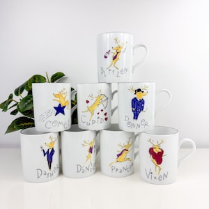 Puede incluir: Ocho tazas de cerámica blanca con bordes plateados, cada una adornada con un personaje de reno de dibujos animados. Las tazas están etiquetadas con nombres como "Dasher", "Dancer" y "Vixen". Las ilustraciones de renos están en varias poses.