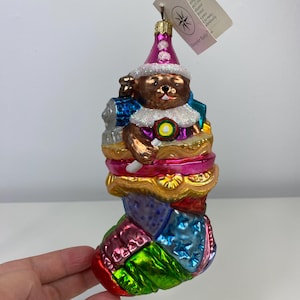 May include: A festive Christmas ornament depicting a bear in a clown costume. The ornament features a pink glittery hat and a patchwork design in shades of blue, green, and red. The bear is holding a small white item.