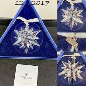 May include: A clear, multi-pointed crystal star ornament featuring a white ribbon and a small metal tag engraved with the year 2017. The ornament is presented in a triangular blue velvet-lined box, accompanied by a white Swarovski warning card.