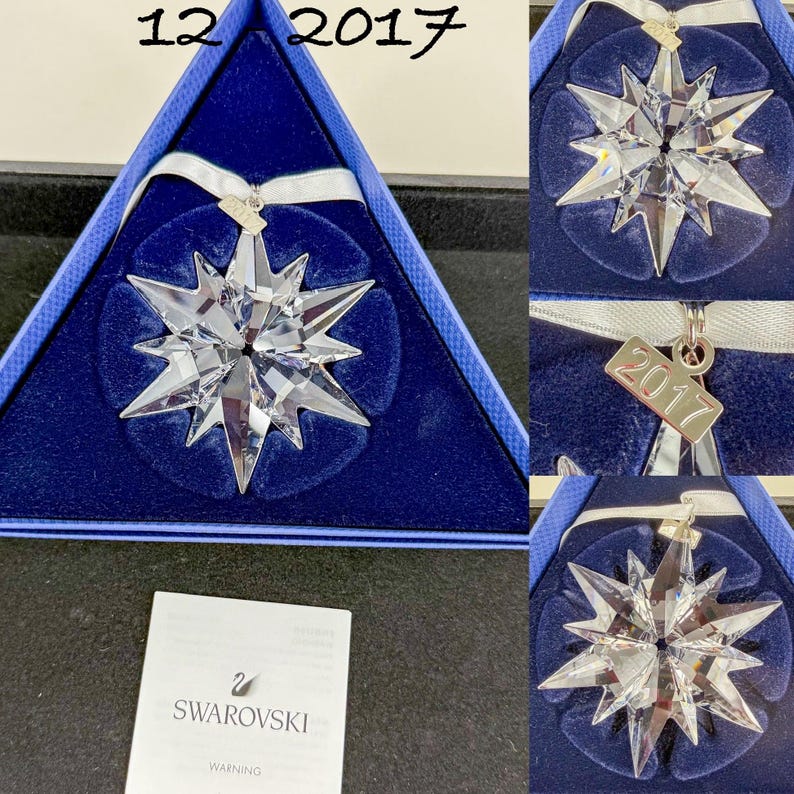 May include: A clear, multi-pointed crystal star ornament featuring a white ribbon and a small metal tag engraved with the year 2017. The ornament is presented in a triangular blue velvet-lined box, accompanied by a white Swarovski warning card.