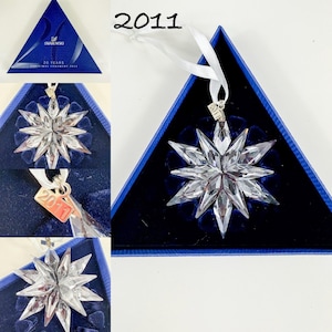 May include: A clear, multi-pointed star-shaped crystal ornament with a white ribbon hanger. A small metal tag attached to the ribbon is engraved with the year 2011. The ornament is presented in a triangular blue box with the Swarovski logo.