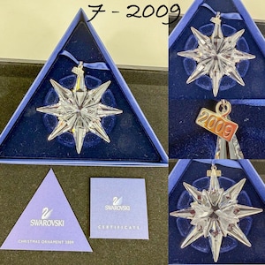 May include: A clear, multi-pointed star-shaped crystal ornament with a silver-toned metal tag engraved with the year 2009. The ornament is presented in a triangular blue box with a blue ribbon and includes a Swarovski certificate of authenticity.