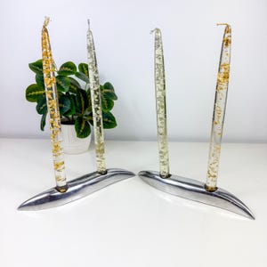 May include: Four clear taper candles embedded with gold and silver metallic flakes, each standing in a sleek, curved, polished silver metal base. The candles are arranged in two pairs, with each pair held by a single elongated metal stand.