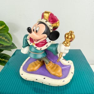 Op de afbeelding: Een beeldje van Mickey Mouse gekleed als koning, met een rode en gouden kroon en een gouden scepter. Het beeld draagt een groene tuniek en een paarse cape, staand op een witte basis met zwarte stippen. Een Disney verzamelobject.