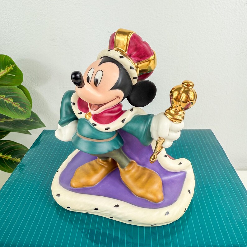Op de afbeelding: Een beeldje van Mickey Mouse gekleed als koning, met een rode en gouden kroon en een gouden scepter. Het beeld draagt een groene tuniek en een paarse cape, staand op een witte basis met zwarte stippen. Een Disney verzamelobject.
