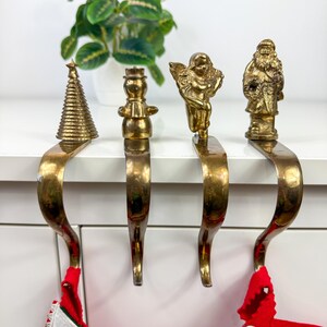 May include: Four brass stocking holders featuring holiday-themed figures: a conical Christmas tree, a snowman, a winged angel, and Santa Claus. Each holder has a curved hook design for hanging stockings on a mantel or shelf edge.