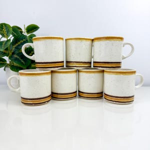 May include: A set of seven ceramic mugs with a speckled cream-colored finish. Each mug features a band of brown and gold stripes near the base and rim. The mugs have a simple, rounded handle.