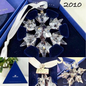 May include: A clear faceted crystal snowflake ornament with a white ribbon hanger and a metal tag engraved with the year 2010. The ornament is presented in a dark blue velvet-lined box with a matching triangular blue box featuring the Swarovski logo.