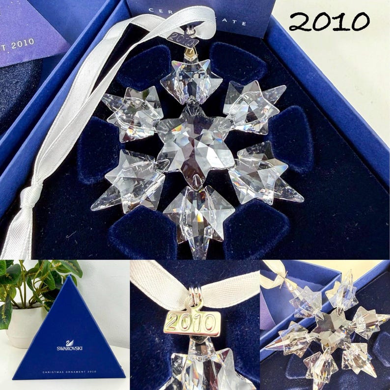 May include: A clear faceted crystal snowflake ornament with a white ribbon hanger and a metal tag engraved with the year 2010. The ornament is presented in a dark blue velvet-lined box with a matching triangular blue box featuring the Swarovski logo.